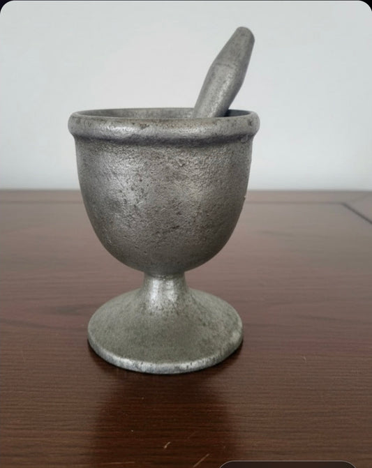 A small antique-style metal mortar and pestle set with a textured, tarnished pewter finish, sitting on a dark wooden tabletop against a plain light gray background.